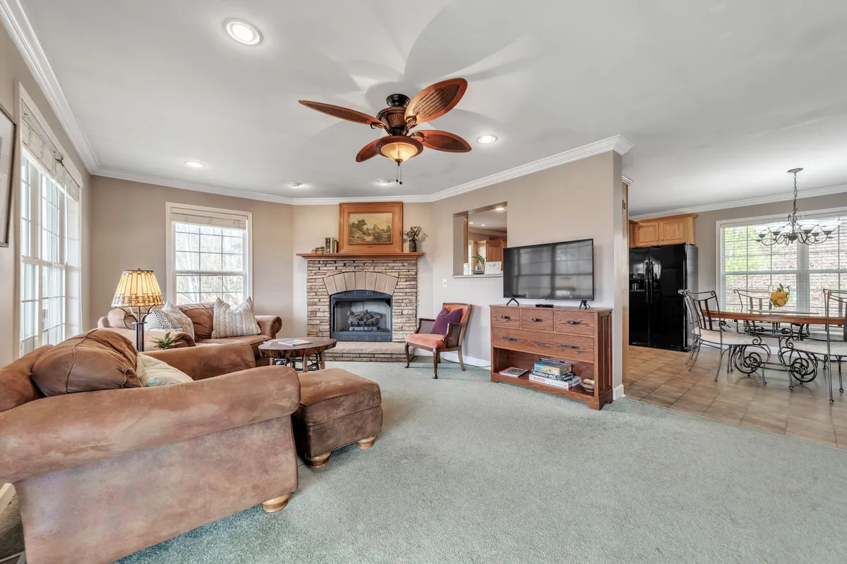 A spacious, open-plan living room featuring comfortable sofas arranged around a cozy fireplace, with a television mounted above and the dining area visible in the background. The space is bathed in natural light, suggesting large windows are present, and the furnishings have a welcoming, homely style.