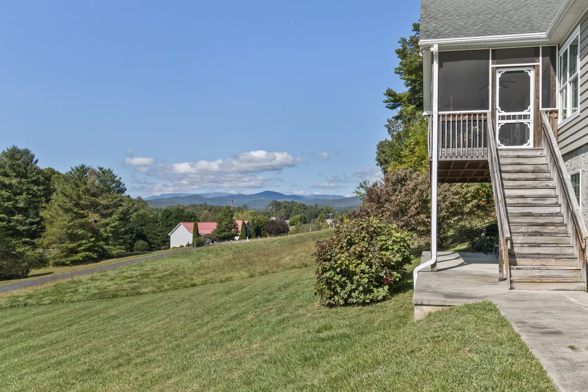 An exterior view looking out over a lush green lawn with a staircase leading to a porch, neighboring houses, and scenic mountain views in the background. The sky suggests a tranquil time for gathering or sunset watching.