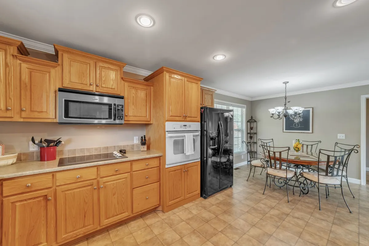 A bright and welcoming dining area with an oak dining table set beneath a chandelier, adjacent to a kitchen with oak cabinetry and black appliances. The space is filled with natural light, evoking a sense of openness and warmth ideal for shared meals and storytelling.
