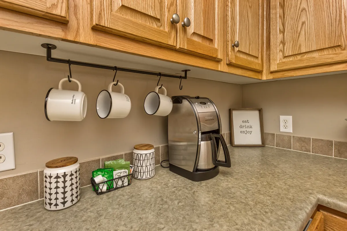 A cozy coffee station setup featuring a sleek coffee maker, a row of hanging mugs, decorative canisters, and a framed quote. The scene evokes the aroma of fresh coffee and the warmth of inviting guests to gather and socialize.