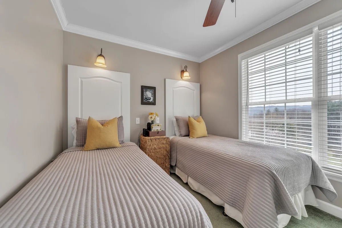 A cozy bedroom with two twin beds featuring bright yellow accent pillows, decorative wall art, and a large window with blinds. The setup encourages shared moments, stories, and relaxation for families or siblings.