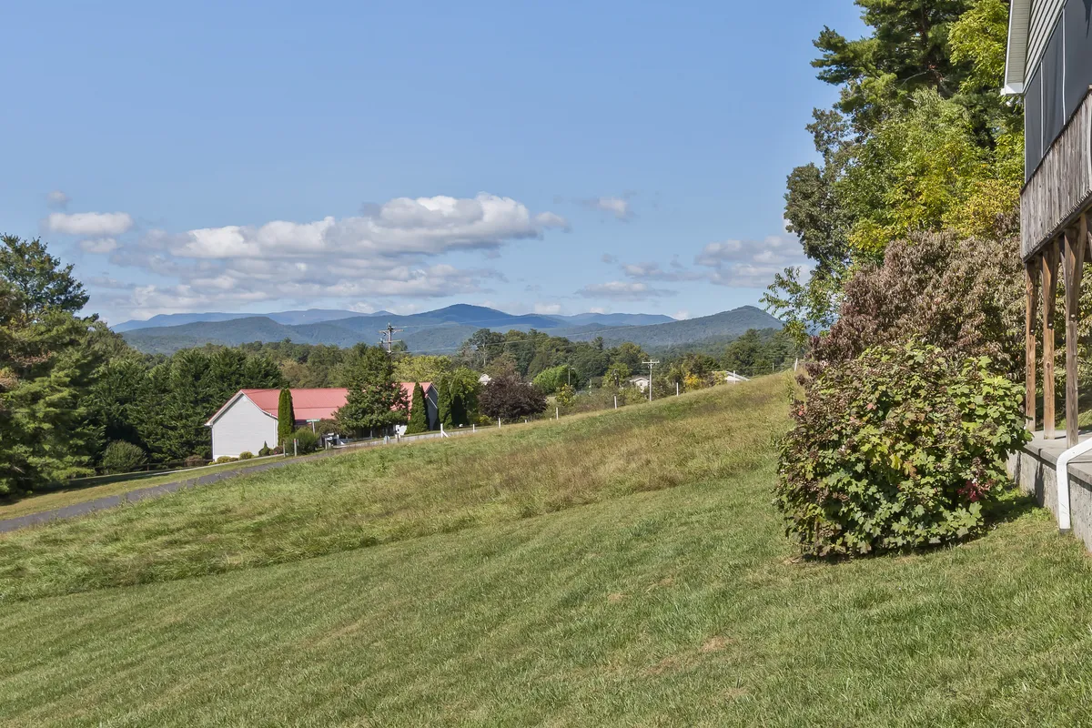 A sweeping view of distant mountains framed by vibrant green grass and a few neighboring houses, the sky glowing with the warm hues of sunset. The open space and tranquil scenery evoke the natural beauty surrounding the property.